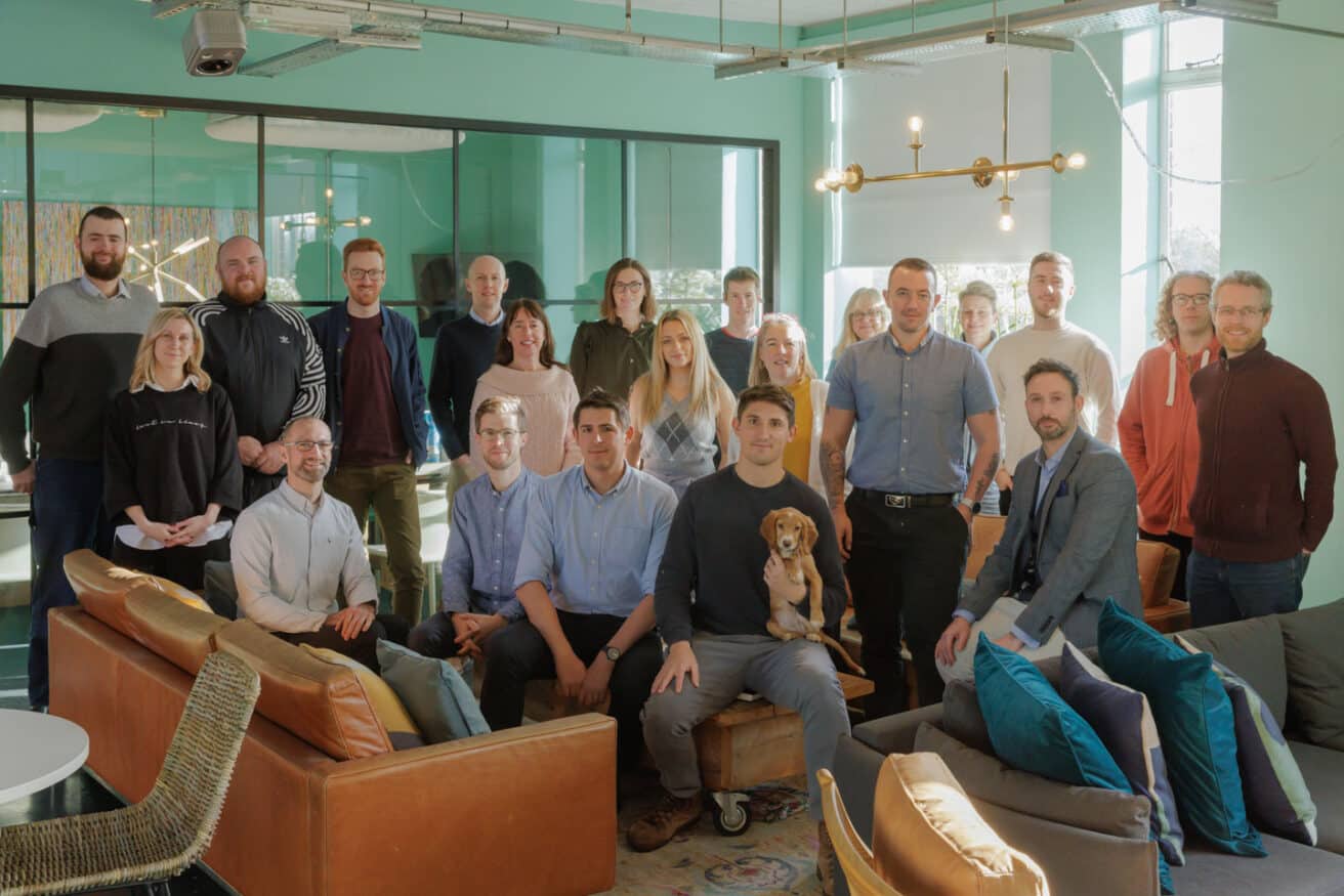 A photo of the CareScribe team in the Bristol office, with everyone gathered around a large sofa and smiling at the camera.