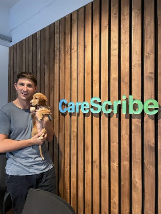 A photo of Rich holding his puppy, Maple, in front of the CareScribe logo on the boardroom wall.