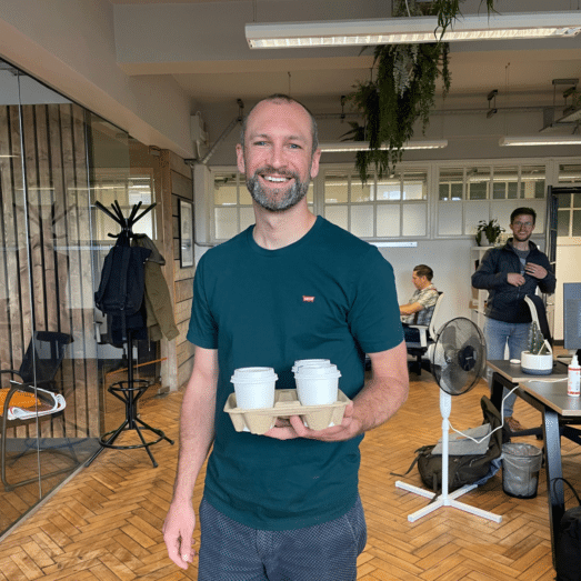 A photo of Paul holding a tray containing three cups of coffee from the local bakery, smiling.