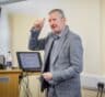 A photo of Gary Taylor who is wearing a grey suit and is putting his thumbs up as he delivers a presentation.