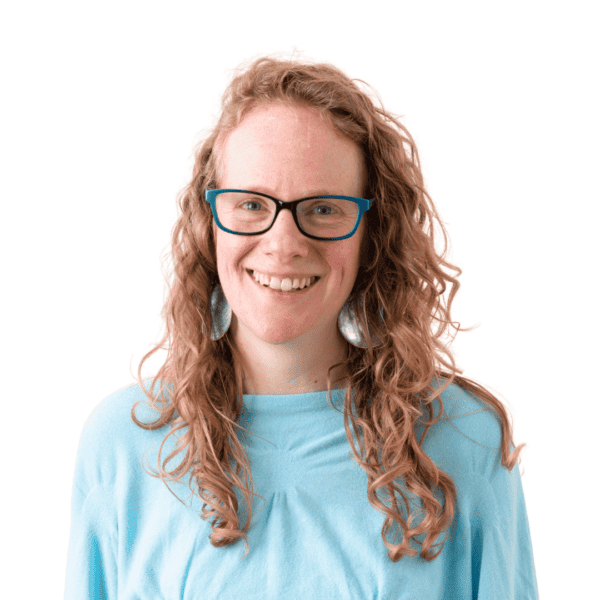A smiling person with curly ginger hair, wearing black-rimmed glasses, large round earrings, and a light blue top, stands against a plain white background—celebrating neurodiversity and mental health awareness in February 2024.