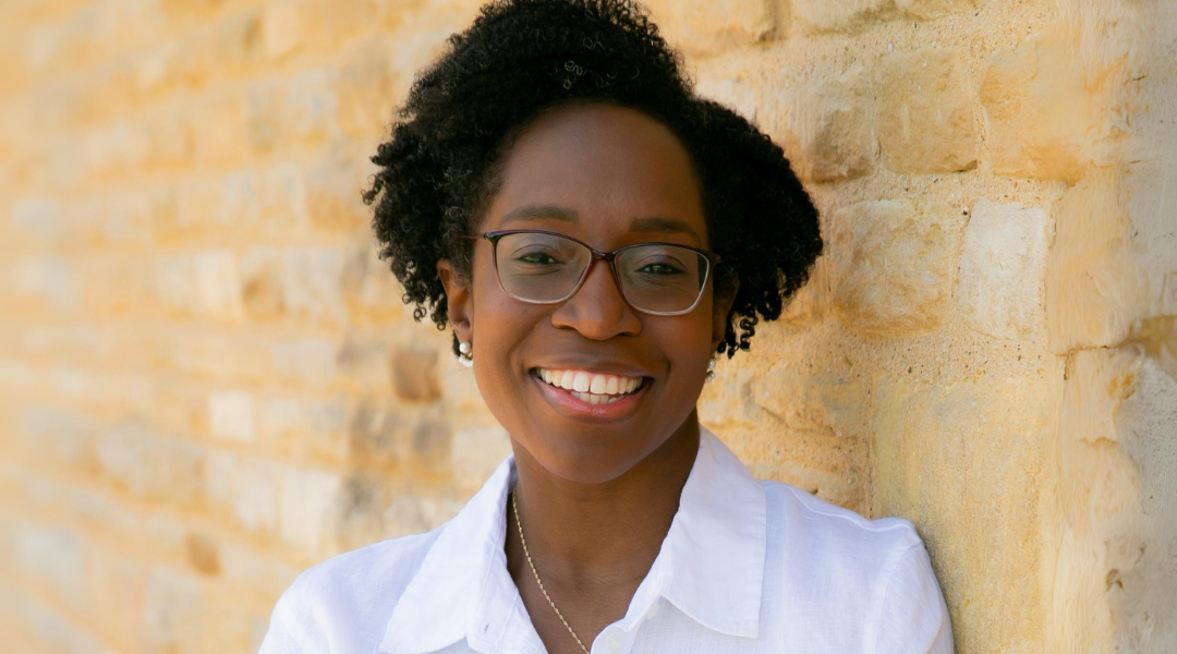 Danielle is a black woman, wearing a white shirt and glasses. She is leaning against a yellow-stone wall and smiling at the camera.