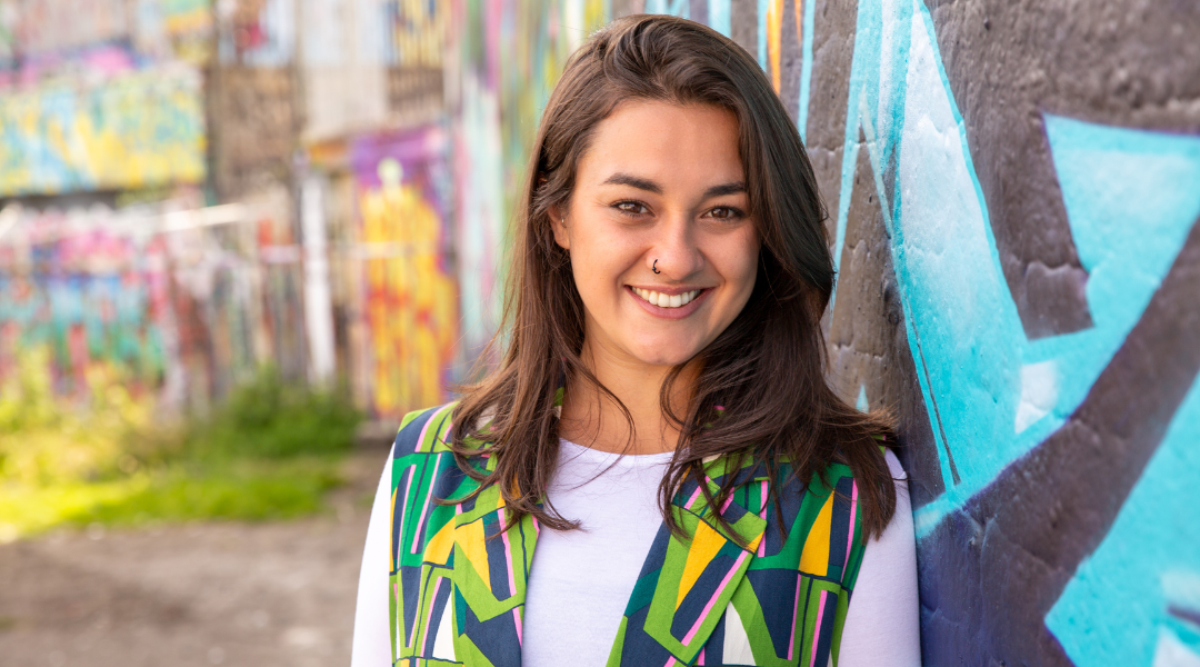 Devon has shoulder-length brown hair with brown eyes and a nose piercing. She is wearing a bold print jacket over a white top and is leaning against a wall with colourful graffiti and smiling at the camera.