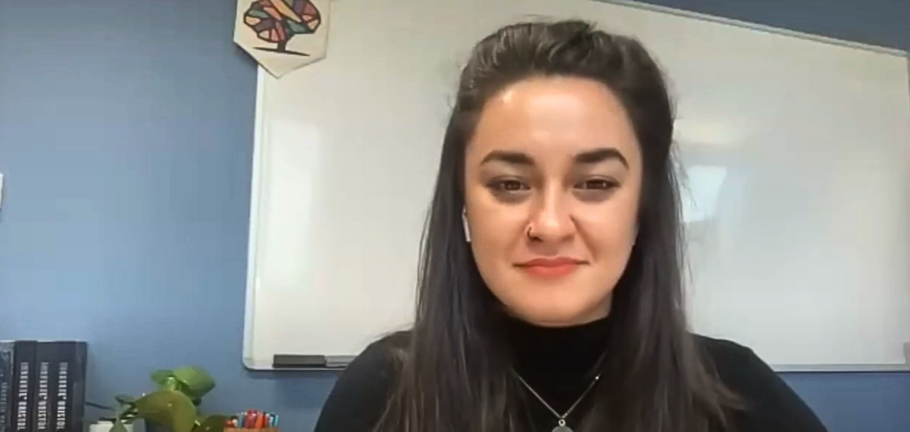 Screenshot of Devon Lowndes during the live webinar. Devon has long brown hair and is wearing a black top and seated in front of a whiteboard.