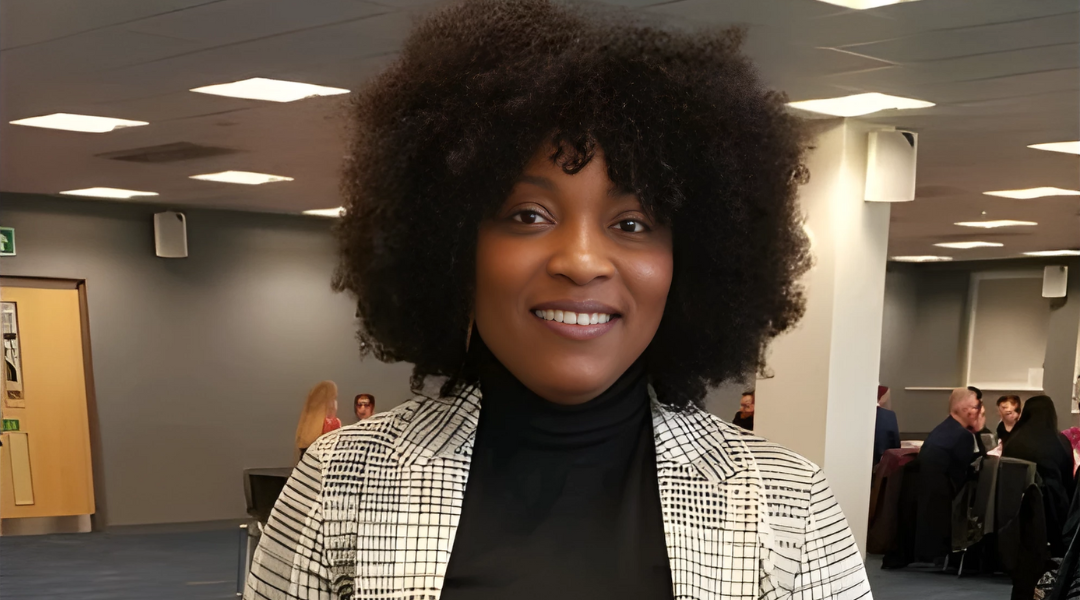Elizabeth Takyi is standing in an office environment wearing a high-neck black top with a black and white blazer over the top. Elizabeth is smiling at the camera.