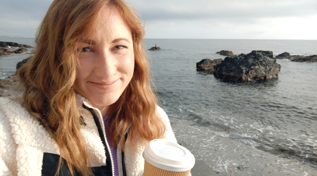 Hannah has medium length hair and is holding a cup of coffee on the beach