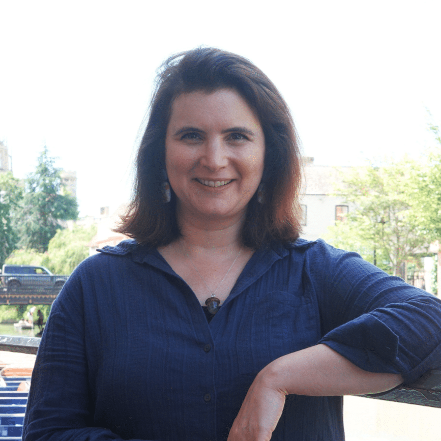 Deborah is standing outside leaning on a railing with dark brow hair and a dark blue top.