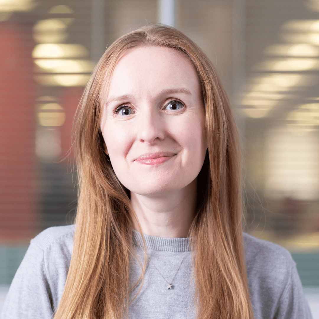 Jen has long hair and is smiling at the camera. She is wearing a grey jumper and in an office setting.