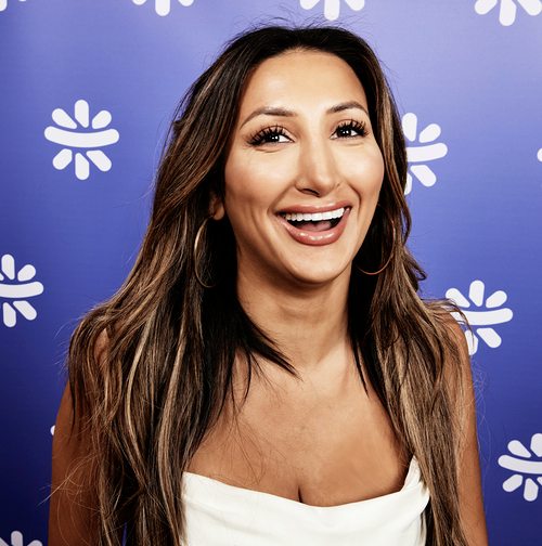 Noor has long brown hair, and dark eyes. She is smiling at the camera in front of a purple background wall with a repeated logo from Not On The High Street.