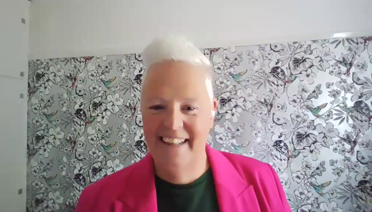 Screenshot taken during the live webinar where Suzi is speaking from a black and white wallpapered room wearing a pink blazer and a black top.