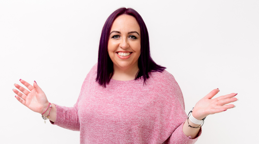 Jodie has deep purple hair and is wearing a pink speckled jumper. She has her arms raised and is smiling at the camera.