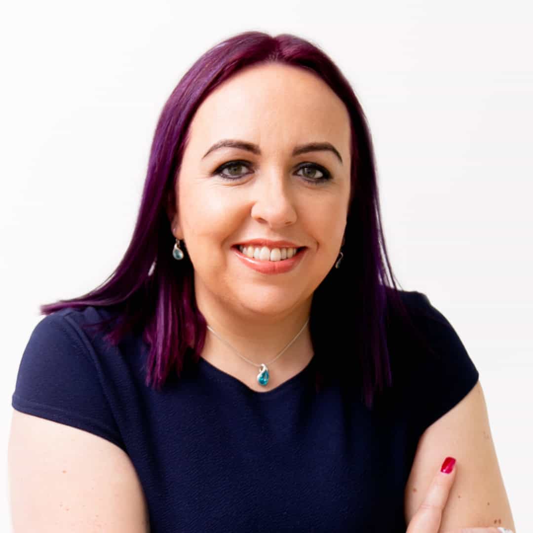 Jodie has deep purple hair and is wearing a navy top with her arms crossed, she is smiling at the camera.