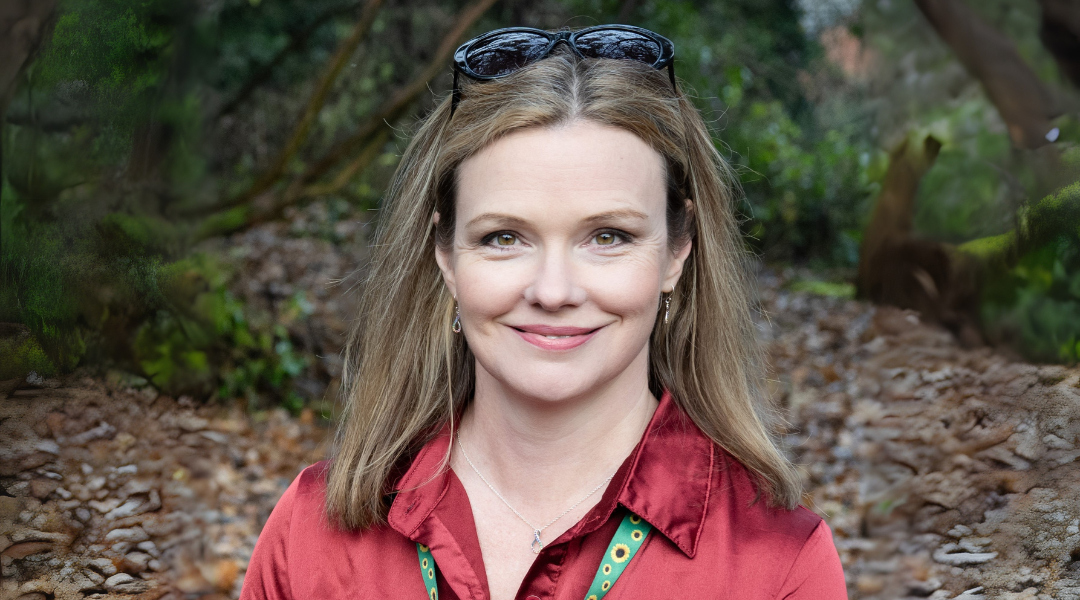 Charlie wears a red shirt, with a sunflower lanyard. She is outside and has black sunglasses on her head. Charlie is smiling at the camera.