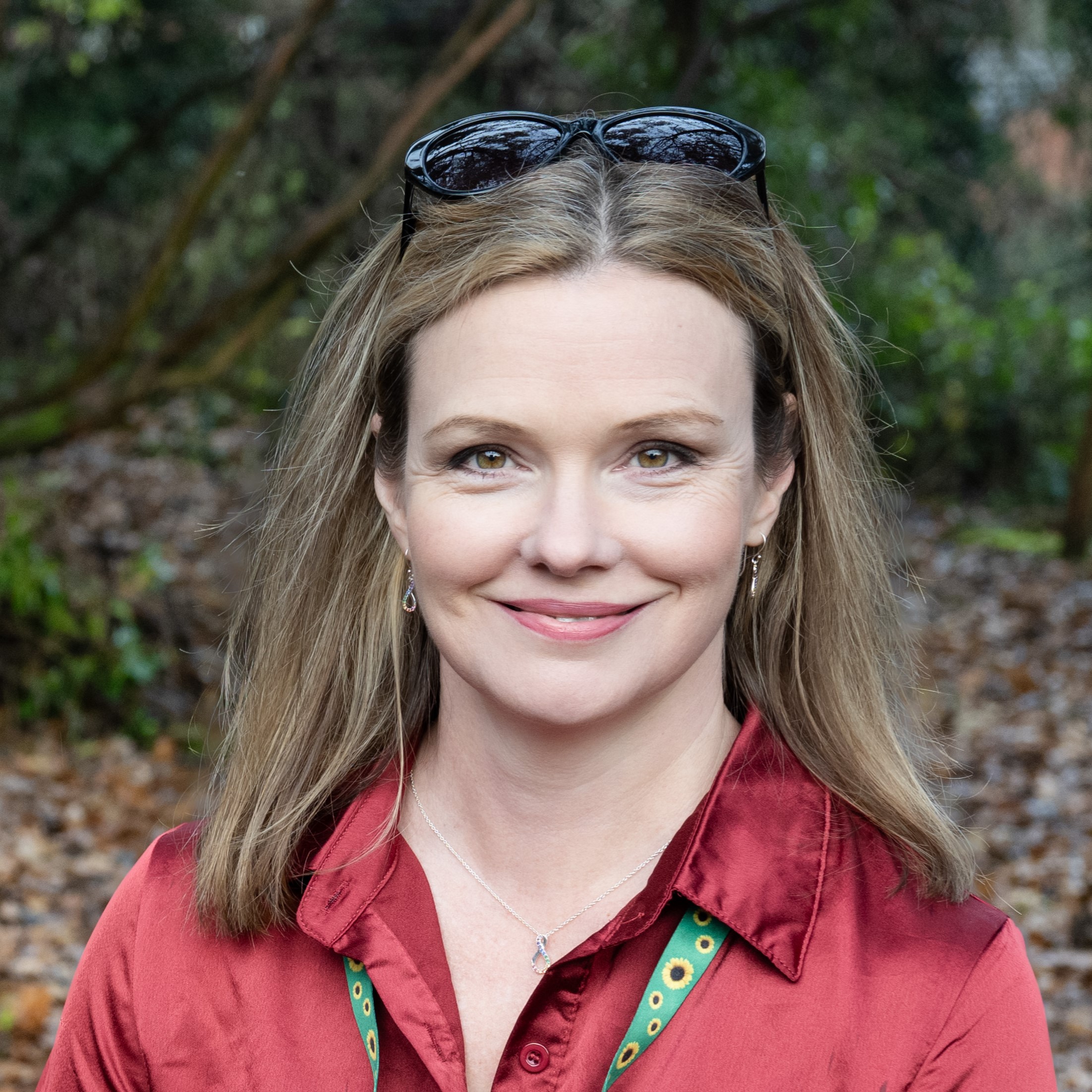 Charlie wears a red shirt, with a sunflower lanyard. She is outside and has black sunglasses on her head. Charlie is smiling at the camera.