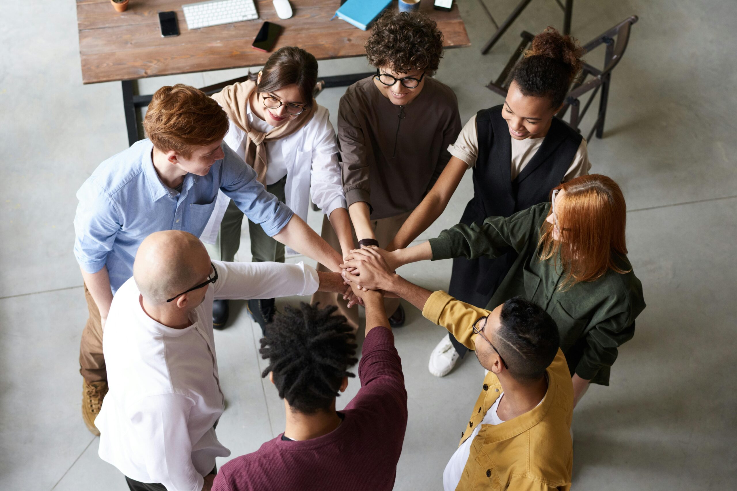A group of eight people stands in a circle with hands stacked together in the centre, embodying collaboration and unity, in a modern office setting with a wooden table and computers in the background.