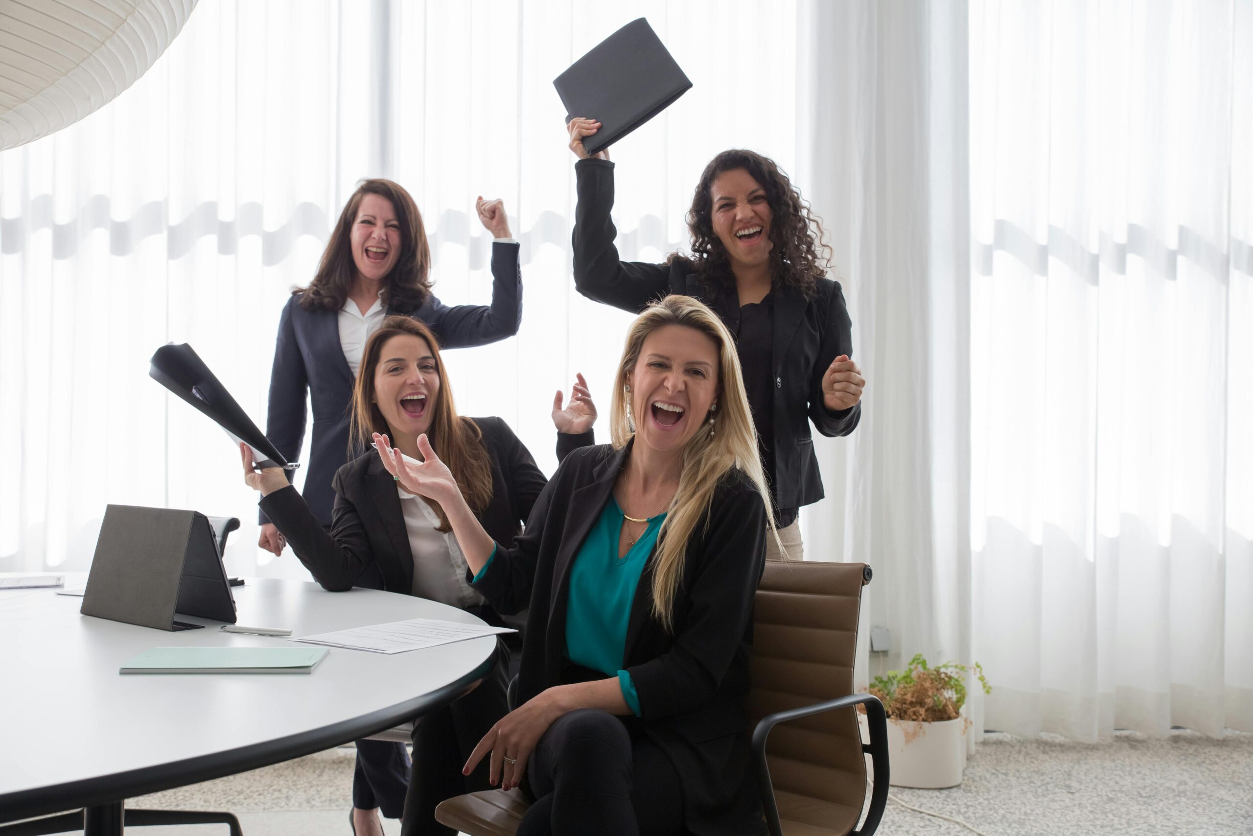 Four women in business attire celebrate together in a bright office, smiling and raising their arms and folders in excitement—showing how self-motivation can help you get unstuck at work and achieve success together.