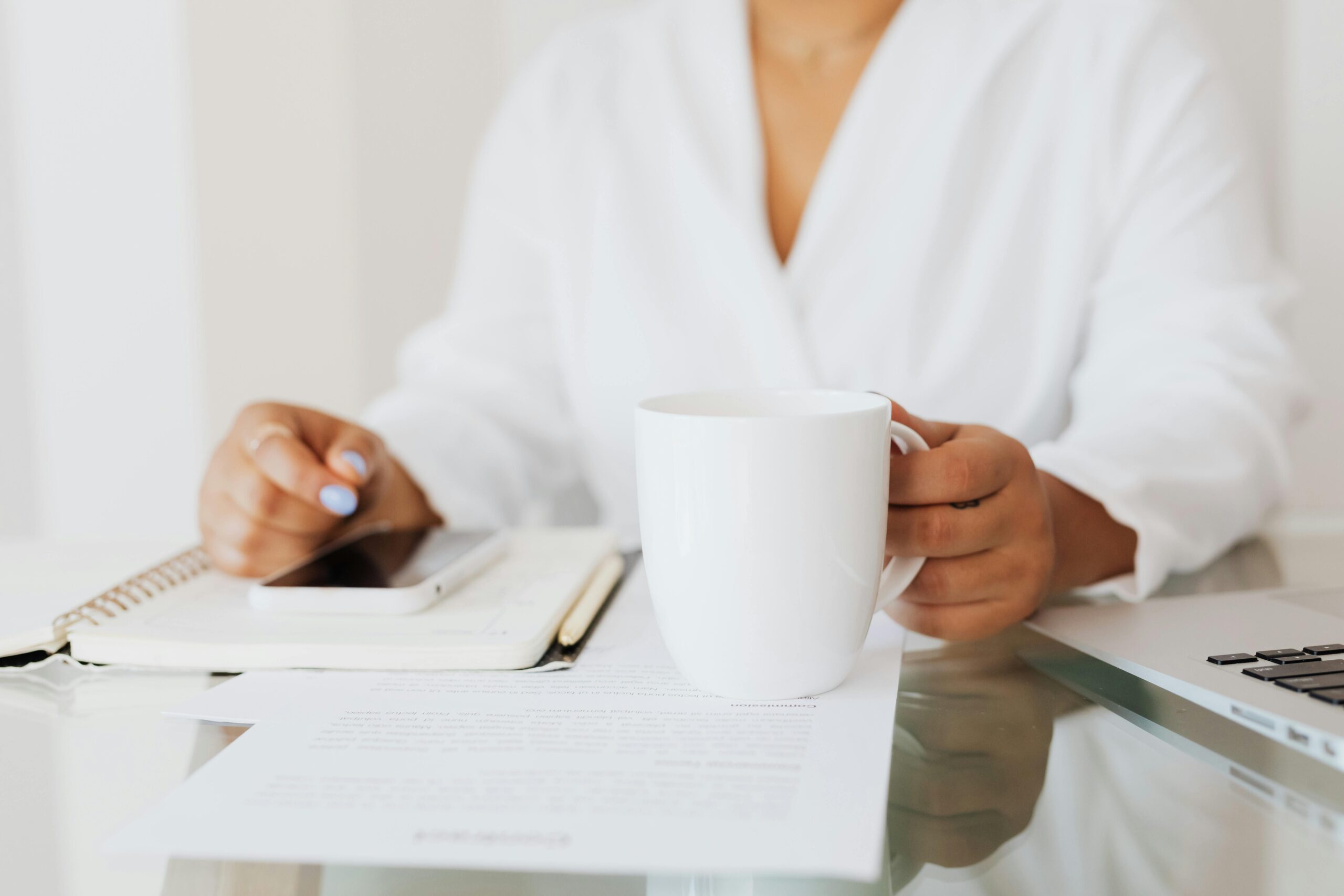 A person in a white shirt sits at a desk holding a white mug, surrounded by an open notebook, mobile phone, and papers—creating a productive morning routine to make your day start off right in a work or study environment.