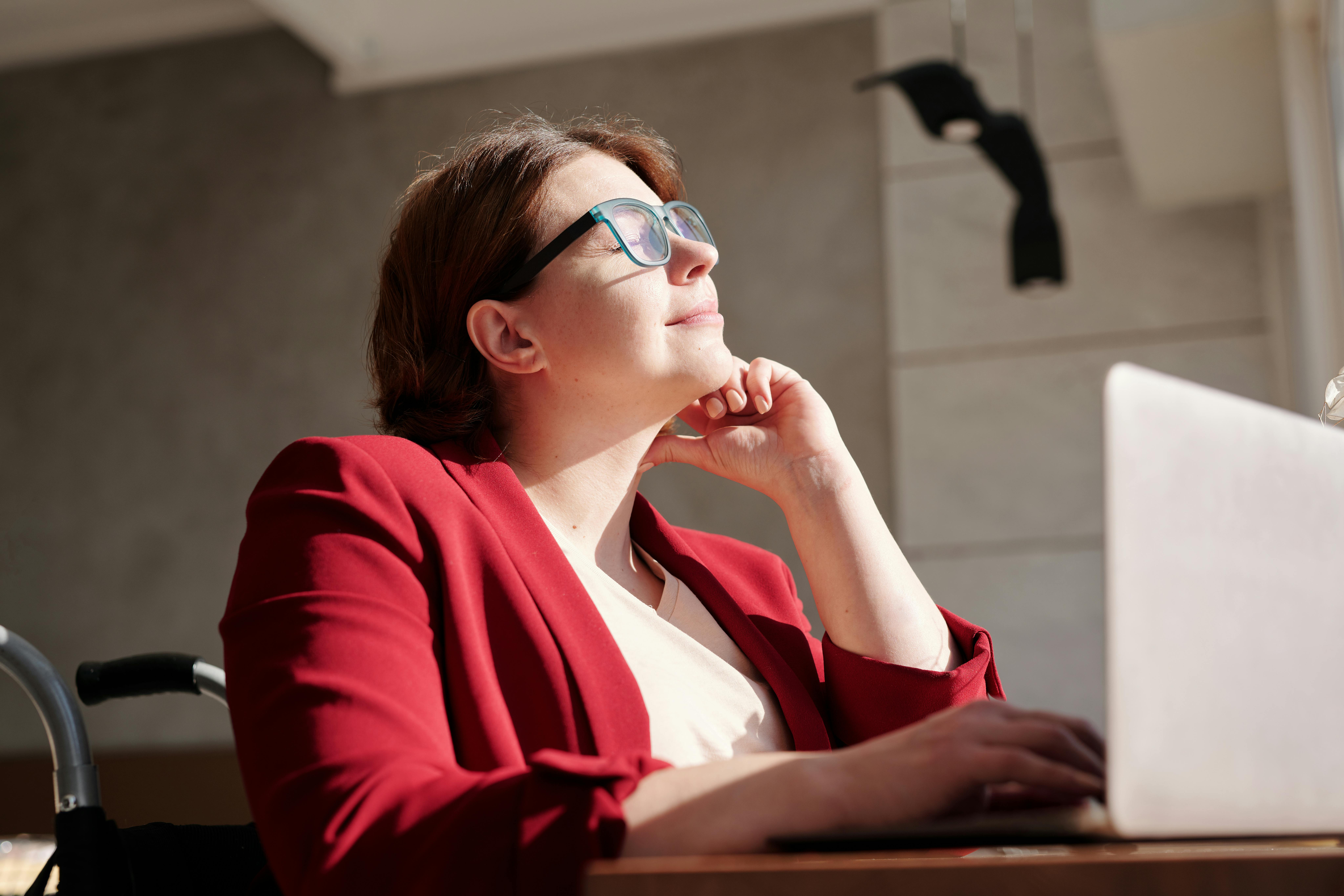 A woman wearing glasses and a red blazer sits at a desk with a laptop, looking up and smiling thoughtfully as sunlight shines on her face, embodying resilience at work.