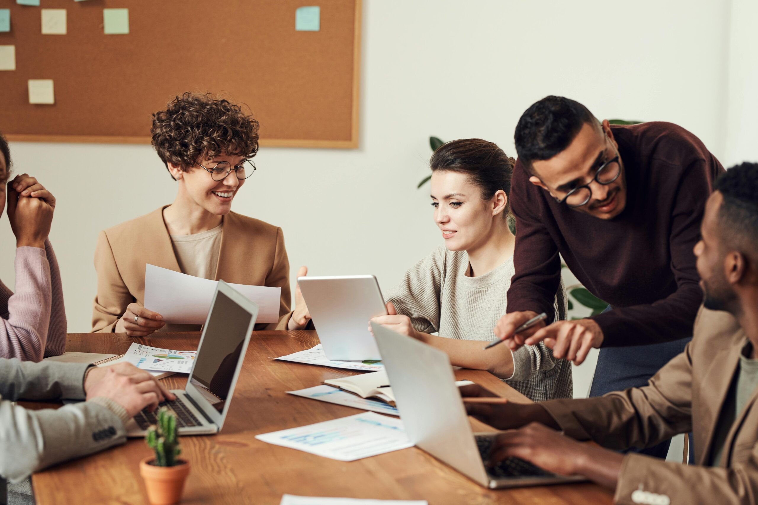 A group of five people sit and stand around a wooden table with laptops, papers, and charts, actively discussing and collaborating in an office setting, showcasing top teamwork skills to succeed, with a noticeboard in the background.