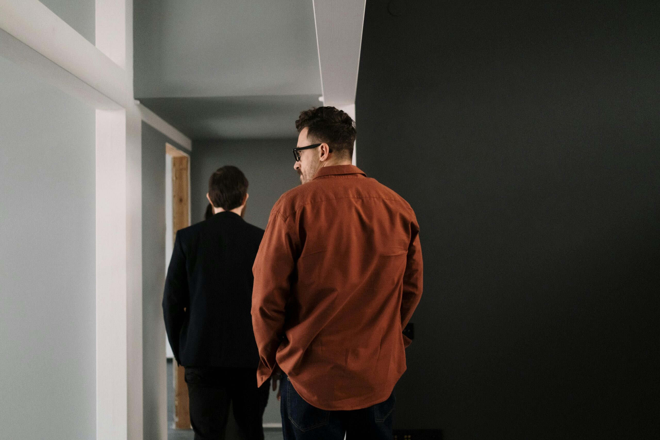 Two men walk down a workplace corridor with white and dark grey walls. The man in front wears a dark suit, while the man behind, in glasses and a rust-coloured shirt, has his hands in his pockets. Both are seen from behind.