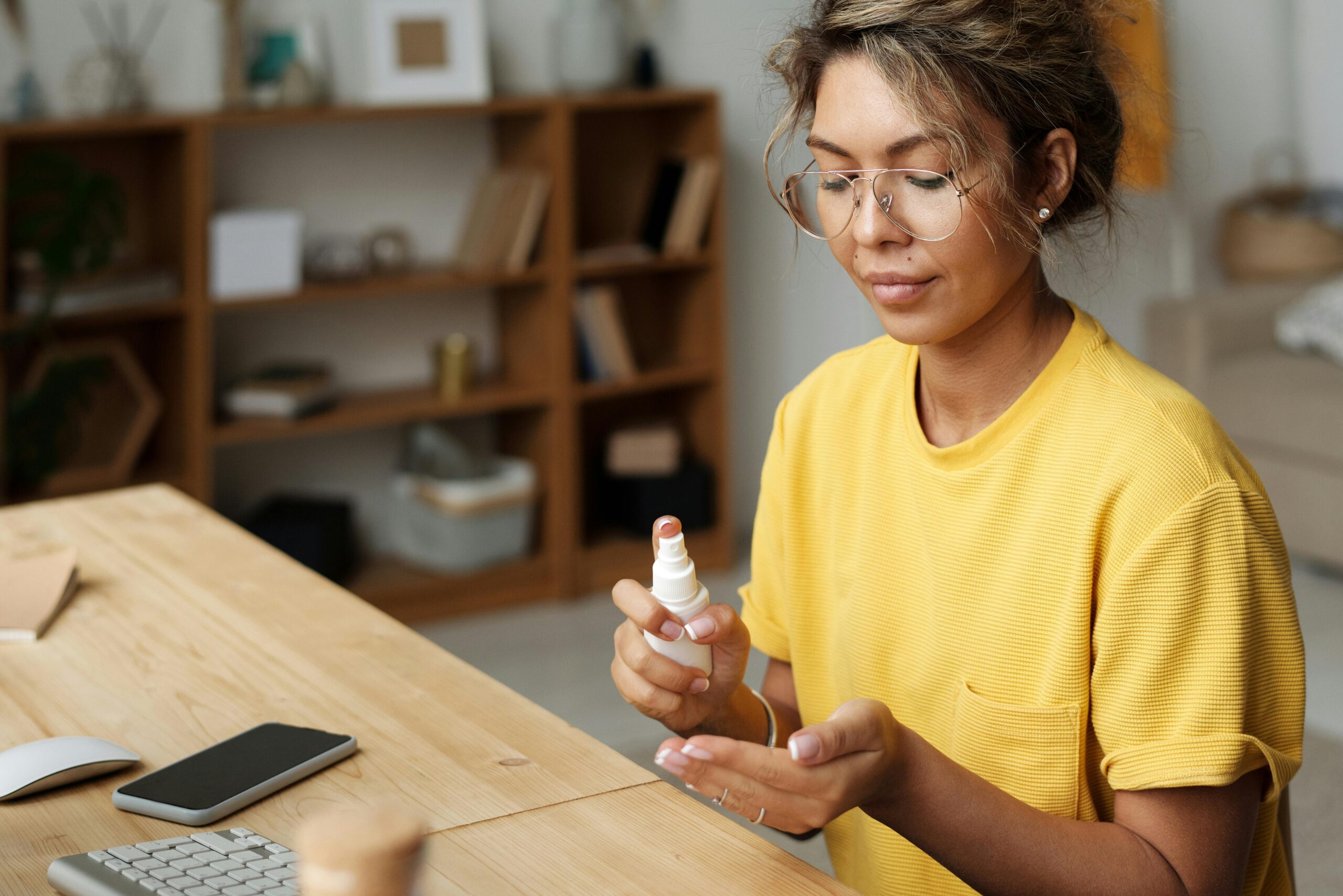 A woman wearing glasses and a yellow shirt sits at a wooden desk, dispensing sanitiser onto her hands—a mindful habit for those seeking OCD support. A mobile phone, keyboard, and papers are on the desk; shelves with books and decor fill the background.