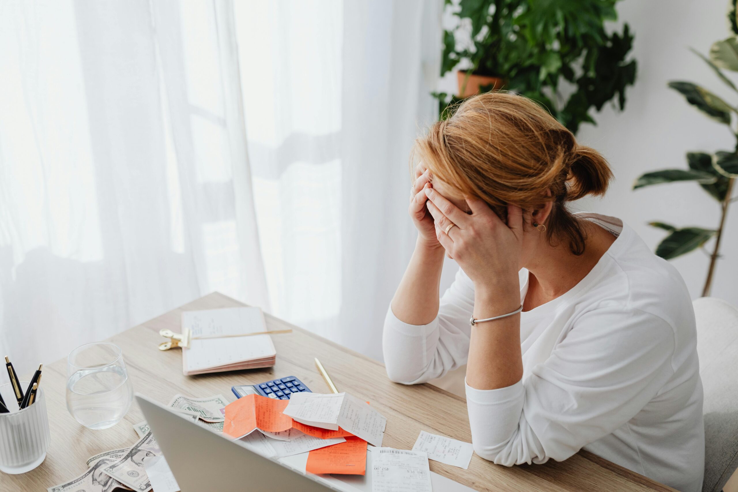 A woman sits at a desk with her head in her hands, surrounded by bills and receipts, appearing stressed as she considers her rights to maximum time off or stress leave UK. A laptop, water glass, and plants are visible in the bright room.