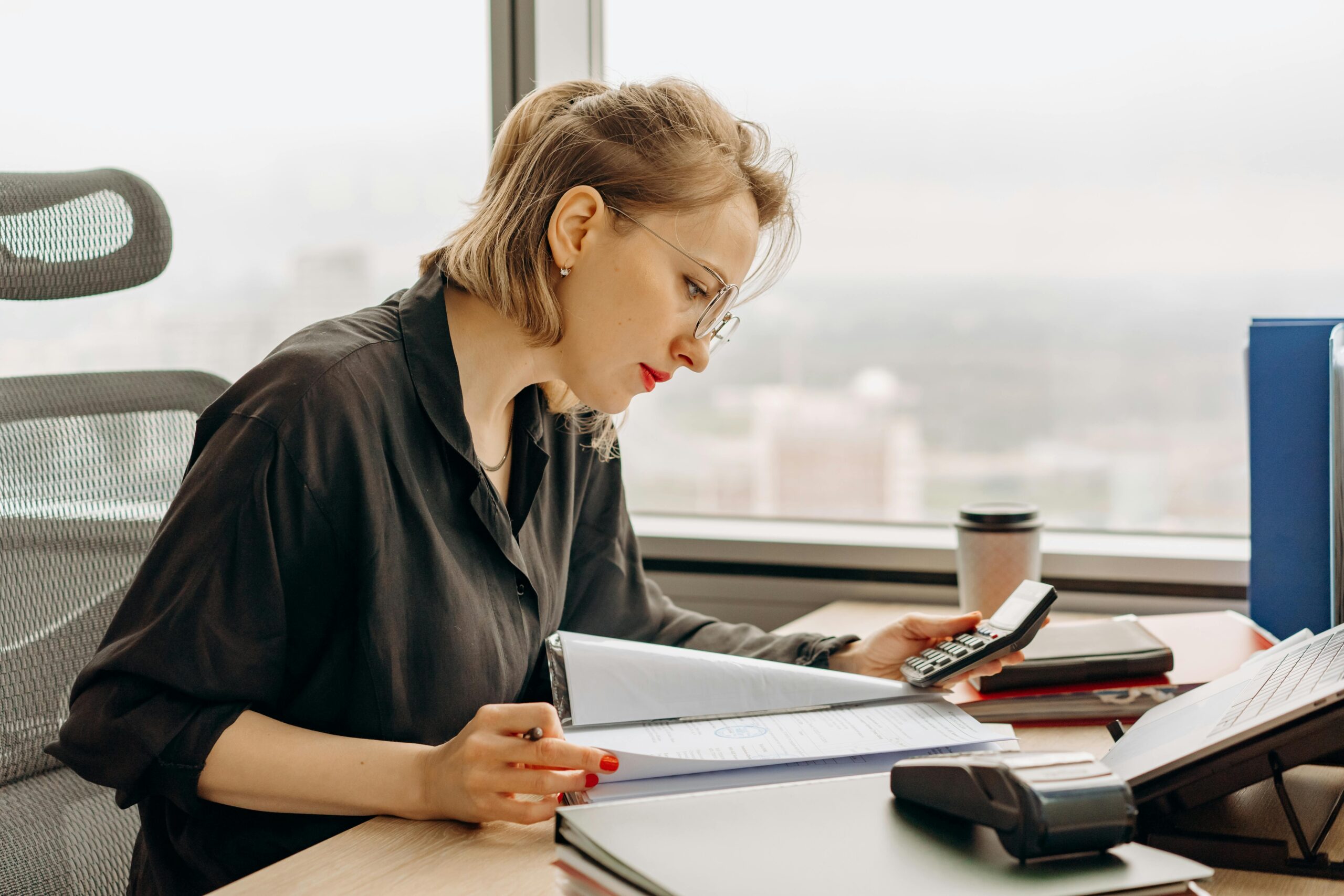 A woman with glasses sits at a desk by a large window, looking at documents in a folder while using a calculator—showcasing strong organisational skills. A computer keyboard, coffee cup, and office supplies are neatly arranged nearby.